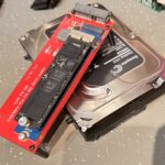 Na zdjęciu widoczny jest dysk twardy Seagate oraz adapter do dysk&oacute;w SSD M.2 NVMe. Adapter leży na tradycyjnym dysku twardym, co sugeruje przygotowanie do analizy danych lub transferu pomiędzy r&oacute;żnymi rodzajami nośnik&oacute;w. Całość jest umieszczona na powierzchni roboczej, prawdopodobnie w laboratorium technicznym.